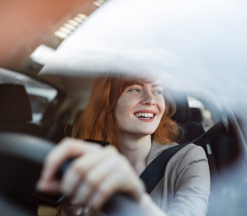 Beautiful smiling young redhead woman behind steering wheel driving car.